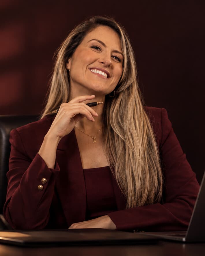 Amanda Leão sorrindo, sentada à mesa do escritório, de blazer bordô, com livros de Direito e uma estátua da Justiça ao fundo.